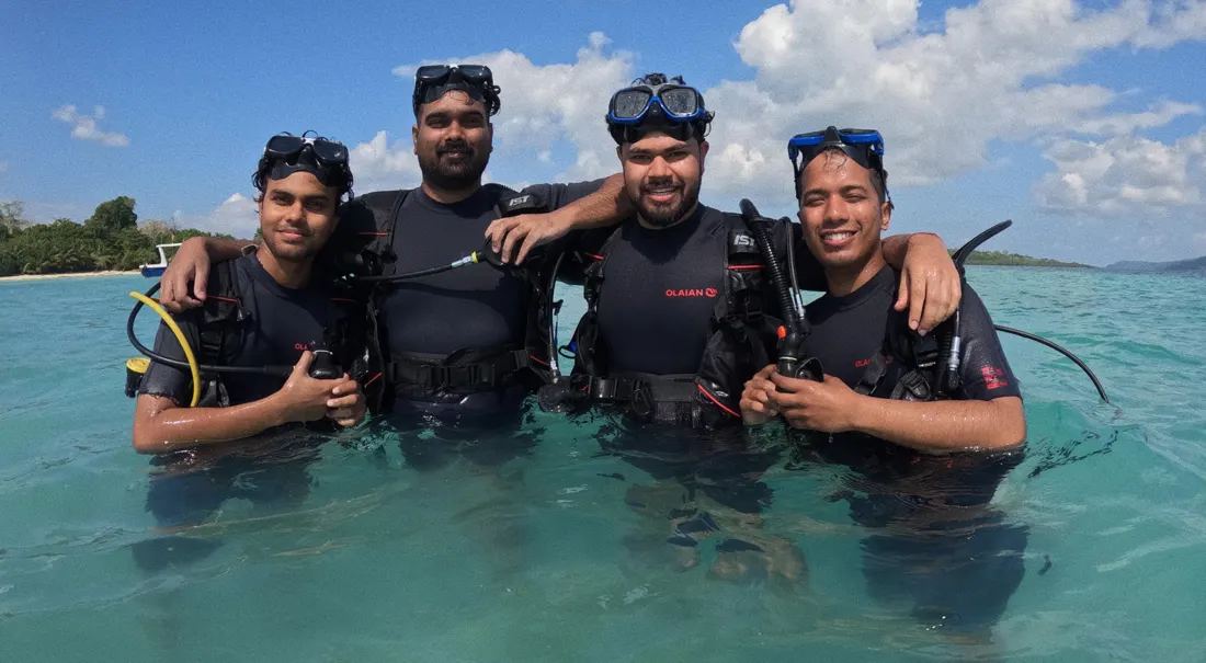 Four divers in wetsuits standing waist-deep in clear water, smiling