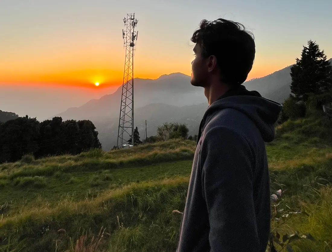 A man in a hoodie looking out over hills as the sun sits on the horizon