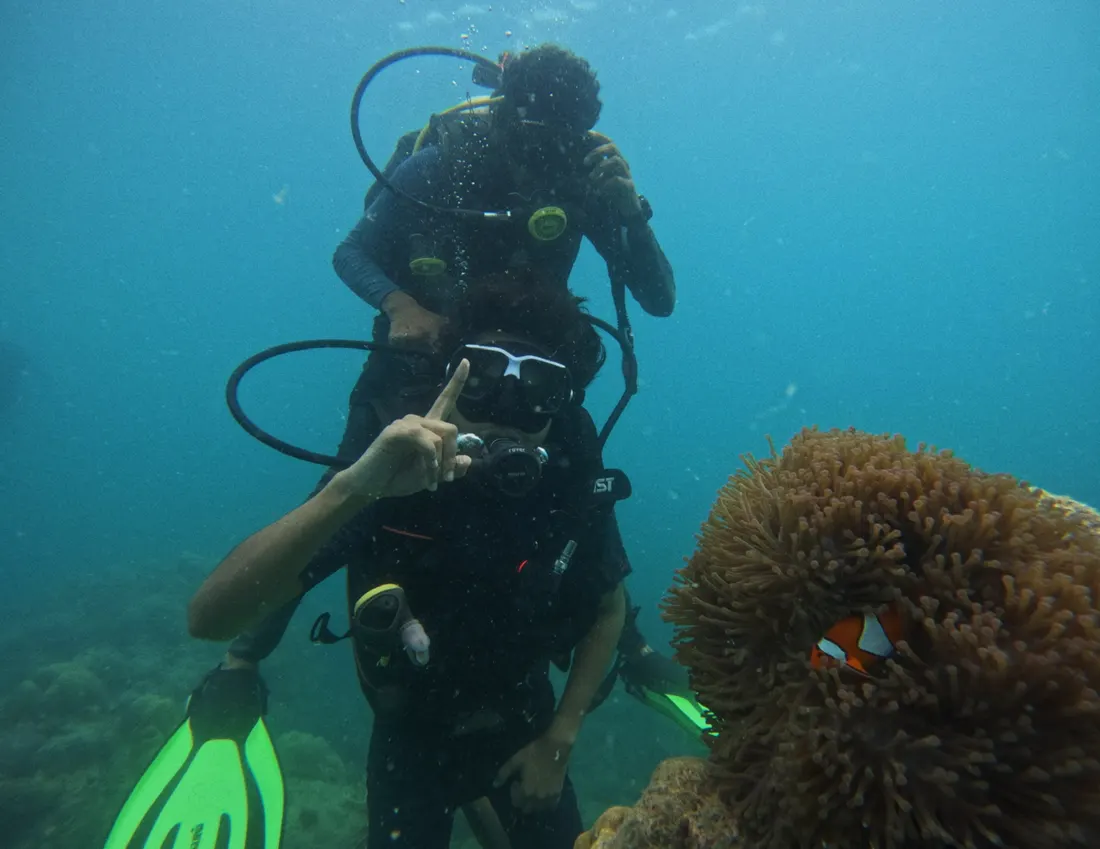 A scuba diver giving a thumbs-up underwater beside a sea anemone with a clownfish