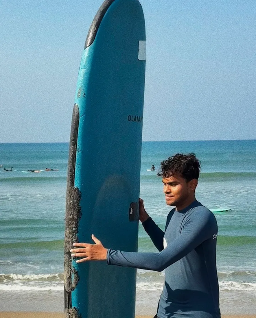 A man in a rash guard standing beside a tall surfboard on the beach