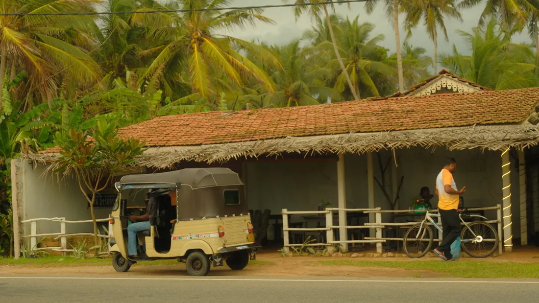 A tuk-tuk parked outside a tiled-roof house under palm trees